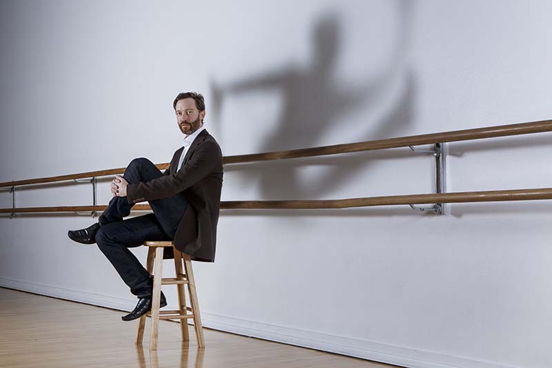 A person sits casually on a wooden stool in a dance studio while a dynamic, dance‑like silhouette is cast dramatically on the wall behind them.