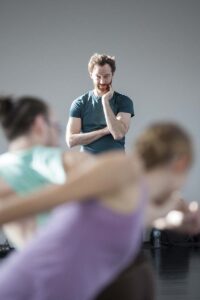 A person standing in a studio observes two individuals in the foreground who are captured in mid‑movement.