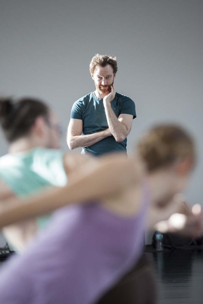 A person standing in a studio observes two individuals in the foreground who are captured in mid‑movement.