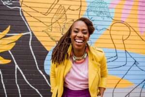 Quynn Johnson is a Black Woman, with brown locs, wearing a yellow motorcycle jacket, pink shirt, and gold, pink, and light blue necklace. She is standing in front of a colorful background with a large smile.