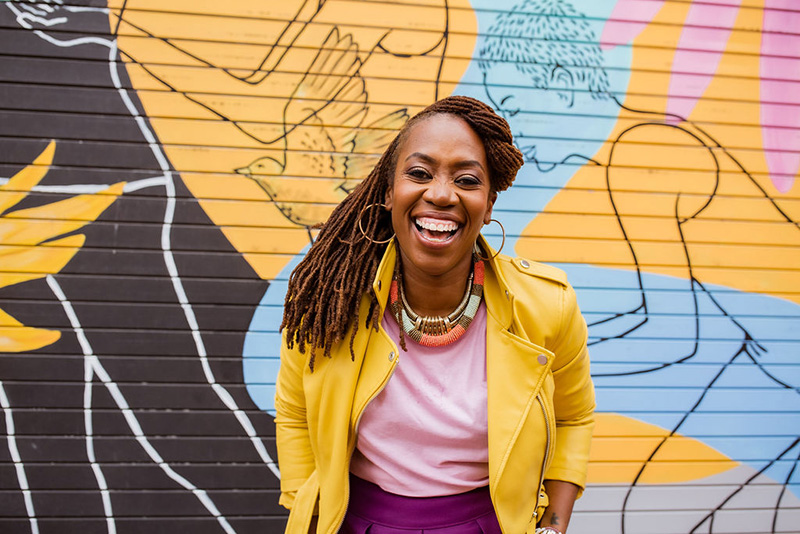 Quynn Johnson is a Black Woman, with brown locs, wearing a yellow motorcycle jacket, pink shirt, and gold, pink, and light blue necklace. She is standing in front of a colorful background with a large smile. 