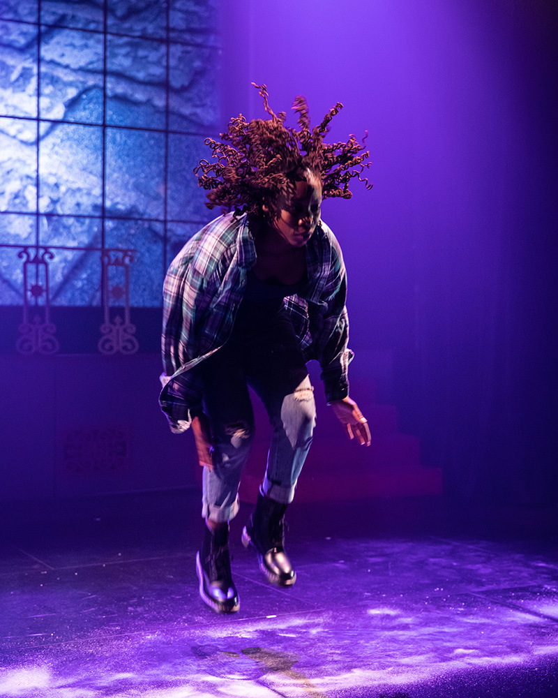 Quynn Johnson, a Black woman with brown locs, is captured mid air while performing on a stage. She is wearing a plaid denim shirt, blue denim jeans, and black boots. The stage is lit with blue and purple lighting. 