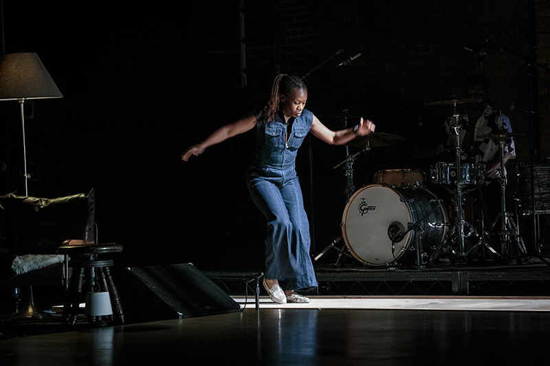 Quynn Johnson is a Black woman dressed in a sleeveless, denim jumpsuit and silver sparkled loafers, dancing in a pool of light. She glances toward the floor as she shifts her weight from one foot to the other. There is a drum set in the background catching some of the light and a collection of living room decorations, including a lamp and a stool, piled in the foreground.
