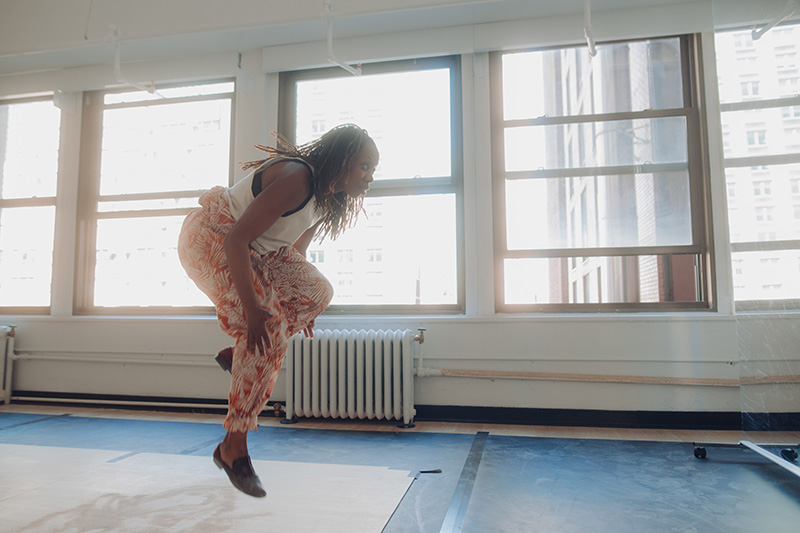 Quynn Johnson, a Black woman dressed in a cream top, orange and cream pants, and black soft shoes, is dancing in a window lined studio. Johnson is caught midair. Her left leg is bent more than her right and her hands reach down towards the sides of her knees. The sunlight is shining bright through the windows. 