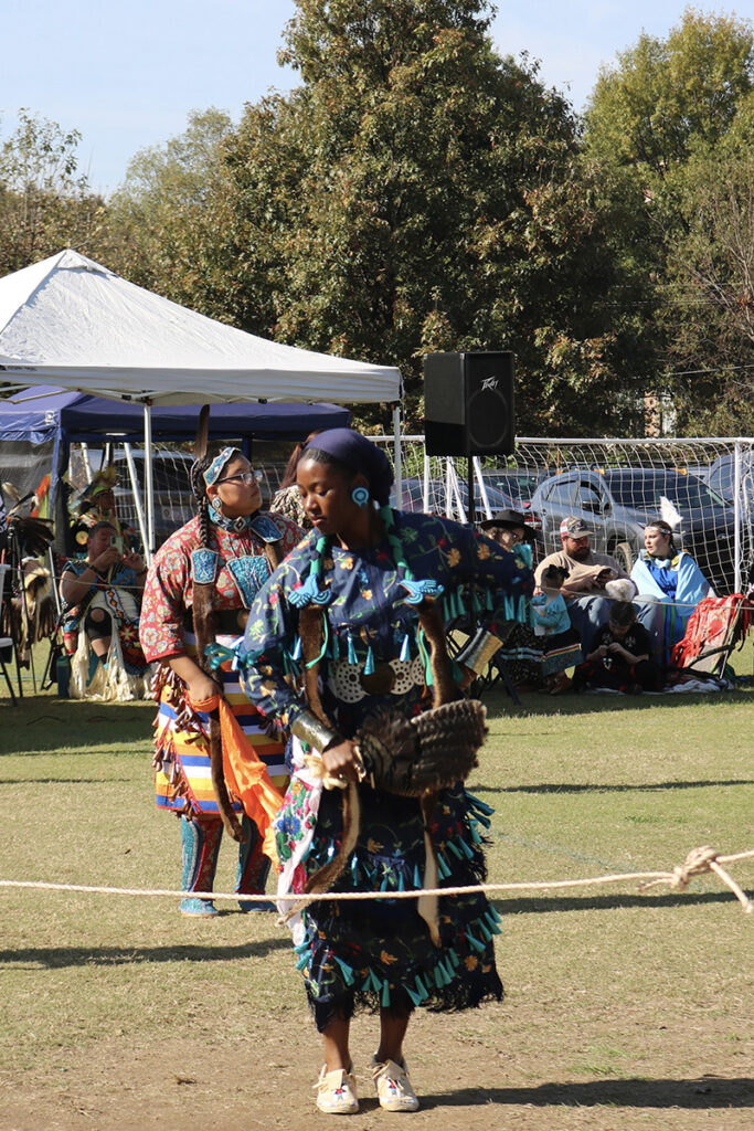 Photo of Umi IMAN dancing at a powwow in Atlanta, Georgia. She is dancing the Jingle Dress Dance, wearing a sacred regalia known as the Jingle Dress. IMAN’s dress, like others of its kind, is adorned with rows of metallic cones that create sound as she moves. The Jingle Dress Dance is a healing dance that originated with the Ojibwe people and is now embraced across many Native communities. Another Jingle Dress dancer appears in the foreground, and the powwow arena is surrounded by spectators.