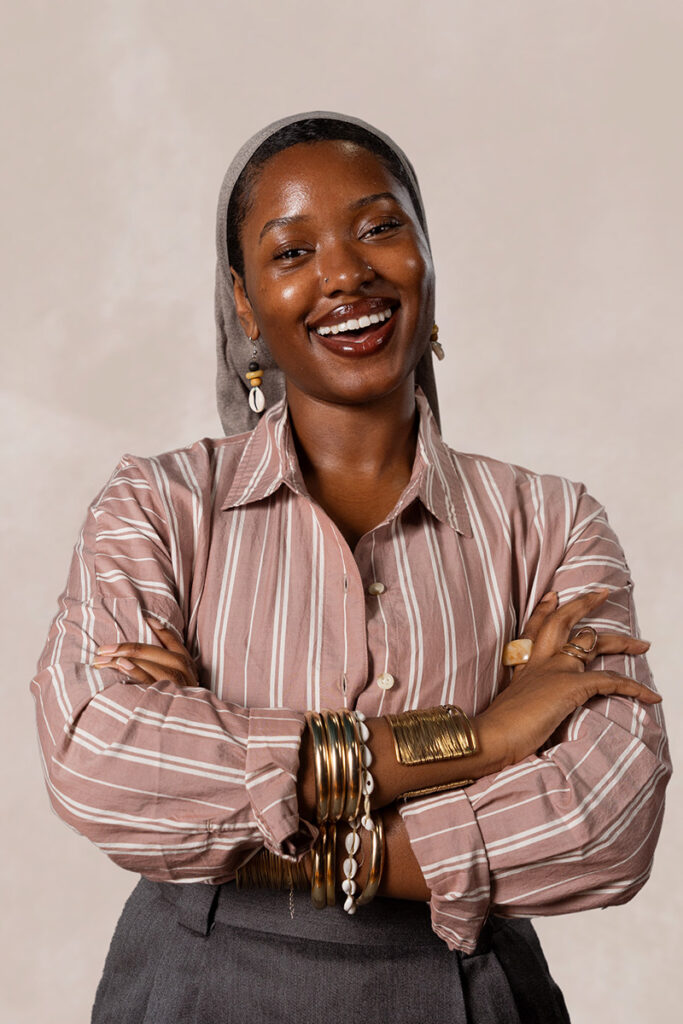 A photo of Umi IMAN. She is smiling and has deep brown skin. She’s wearing a grey hijab with her black hairline slightly showing. She has cowrie shell earrings, deep red lipstick, and a mauve pink button-up shirt with white stripes. She’s also wearing gold and white cowrie shell bracelets, gold and tan rings, and a charcoal grey skirt.