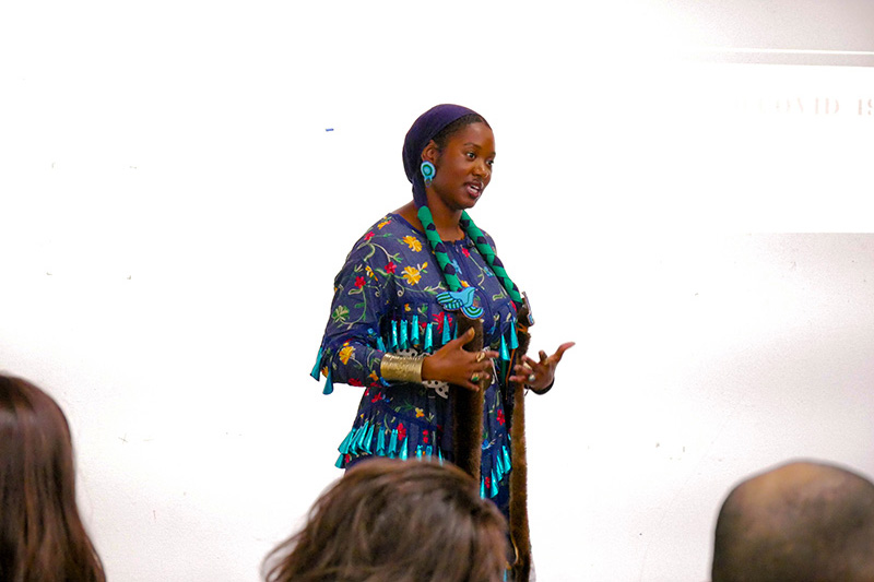 Umi IMAN stands against a white background and wears a sacred regalia known as the Jingle Dress. IMAN’s dress, like others of its kind, is adorned with rows of metallic cones that create sound. She speaks to an audience and gestures with her hands. In the foreground, three heads of the audience members are visible.