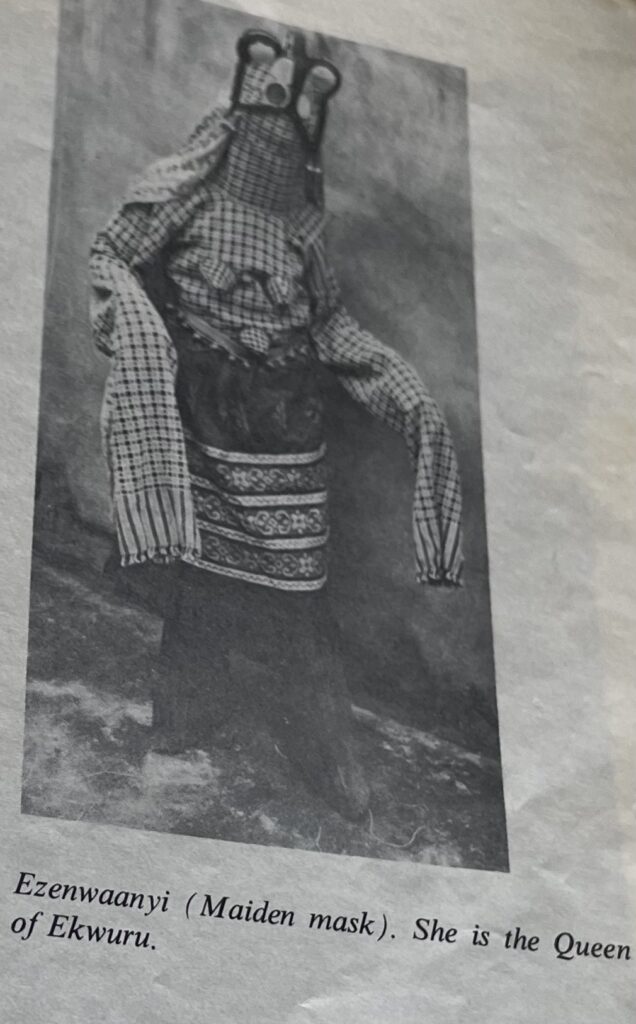 A black-and-white printed photograph shows a costumed ceremonial figure wearing a horned mask, layered patterned garments, and long fabric-covered arms, with a caption below reading “Ezenwanyi (Maiden mask), She is the Queen of Ekwuru.”