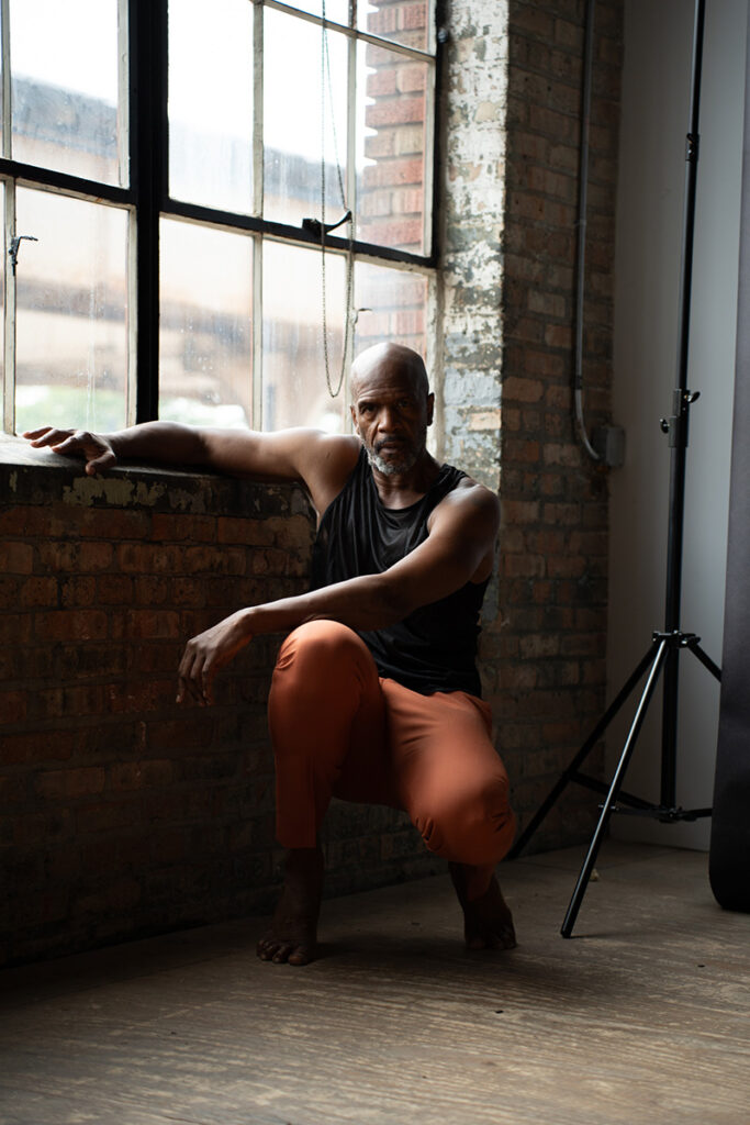 Iega kneels and stares at the camera in a studio next to a window. His arm rests on the sill.