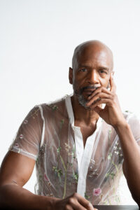 A headshot of Iega wearing a white lacy shirt and holding his chin in one hand.