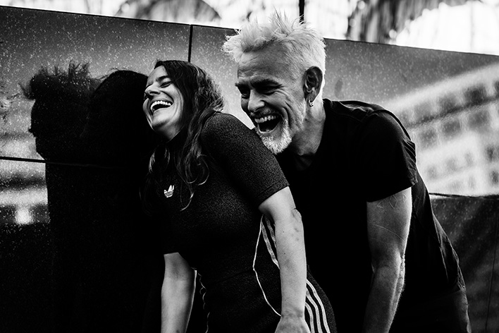 A black-and-white photograph shows Rachael and Jess standing close together against a dark, reflective wall, both leaning forward with open mouths and relaxed postures, captured mid-movement in an urban outdoor setting.