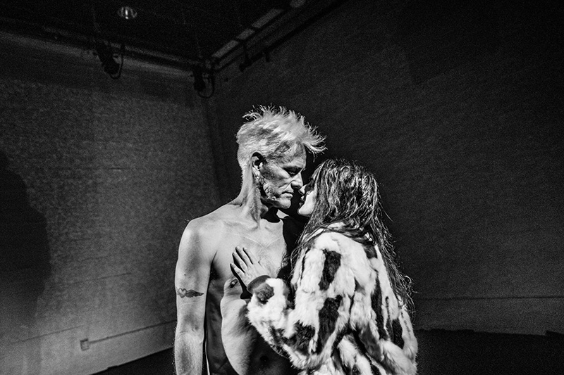 A black-and-white photograph shows Rachael and Jess standing very close together in a dim, sparse indoor space, one naked and the other wearing a patterned fur coat, with their bodies angled toward each other and one hand resting on the other’s chest.