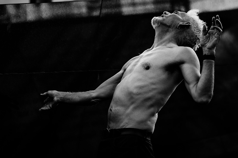 A black-and-white image depicts Jess shirtless standing against a dark backdrop with the torso arched backward, one arm extended downward and the other bent upward near the head, emphasizing the contours of the body through dramatic lighting and contrast.