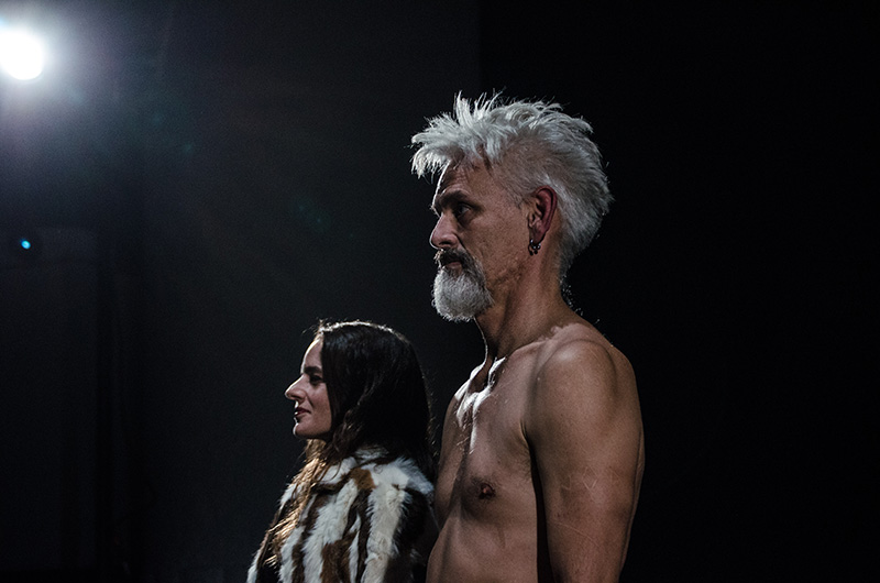 A side-lit photograph shows Rachael and Jess standing in profile against a dark background, Jess naked with short light hair in the foreground and Rachael slightly behind wearing a patterned fur coat, both illuminated by a bright overhead light.