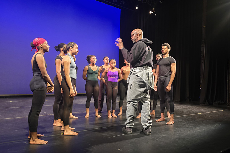 Iega speaks to a group of dancers huddled around him on a stage.