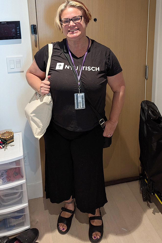 Raven wears a black “NYU TISCH” T-shirt. They stand indoors in front of a wooden door, carrying a tote bag and wearing a lanyard with an ID badge.