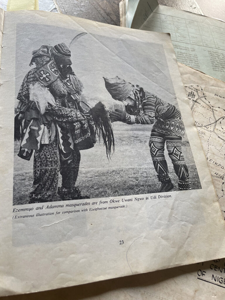A black-and-white printed photograph shows two elaborately costumed masquerade figures standing outdoors, with one figure bowing toward the other while both wear heavily patterned garments and layered ceremonial adornments, accompanied by a caption identifying them as Ezemuọ and Adamma masquerades.