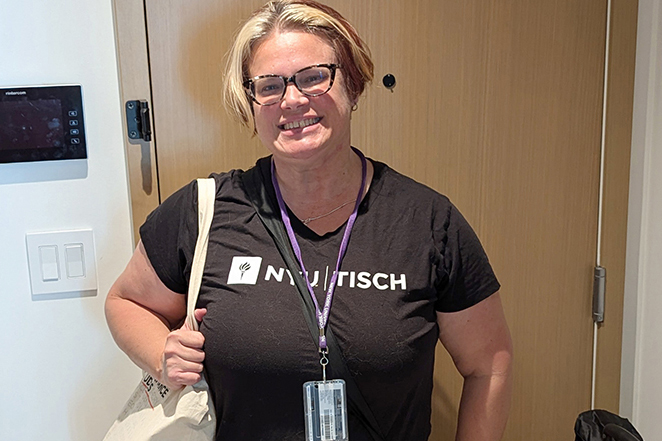 Raven wears a black “NYU TISCH” T-shirt. They stand indoors in front of a wooden door, carrying a tote bag and wearing a lanyard with an ID badge.