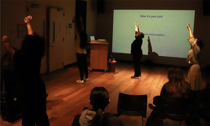 Several people stand on a wooden stage in a dimly lit room raising their arms while a projected slide behind them reads “Now it’s your turn!” as seated audience members watch.