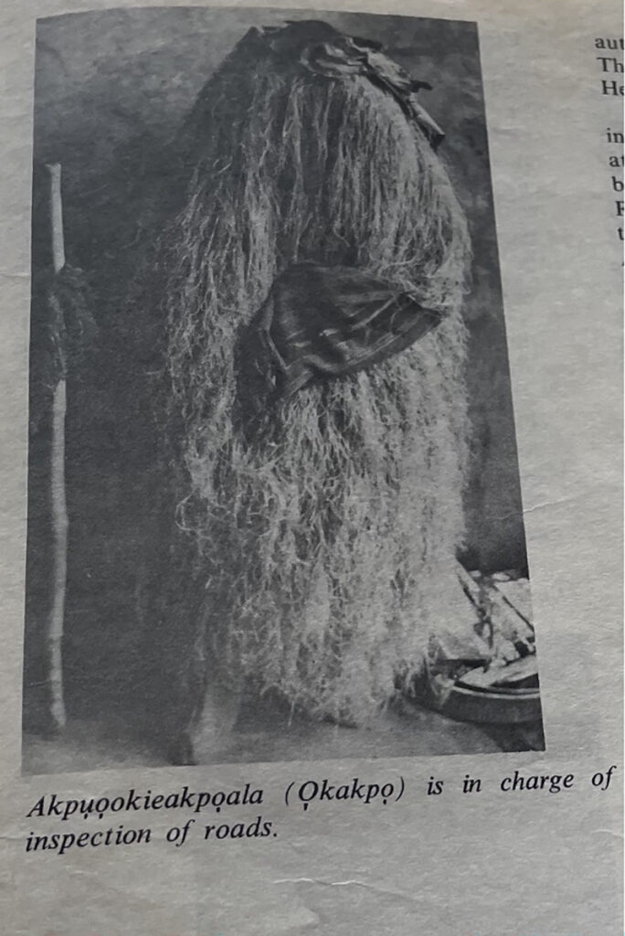 A black-and-white printed photograph shows a masquerade figure completely covered in long raffia fibers holding a staff, with a caption identifying the figure as “Akpuọokiekapụala (Ọkakpọ),” responsible for the inspection of roads.