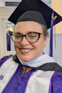 Raven wears glasses and a graduation cap with a purple tassel, posing indoors in a purple gown and white academic hood.