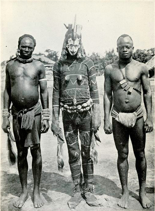 A black-and-white photograph shows three barefoot people standing outdoors side by side, with the central person wearing a full-body patterned costume and mask with horns and adornments, flanked by two minimally dressed individuals wearing necklaces, armbands, and waist cloths.