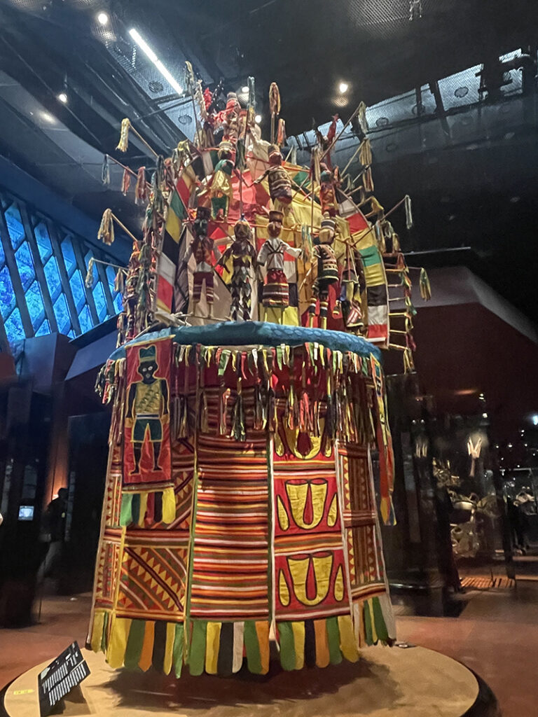 A tall, vividly painted ceremonial structure covered in geometric patterns, hanging ornaments, and small carved figures stands on display inside a museum gallery under focused lighting.
