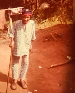 A color photograph shows an elderly person standing barefoot on a reddish dirt path outdoors, leaning on a walking stick and wearing a patterned outfit and cap, with trees and household items visible in the background.