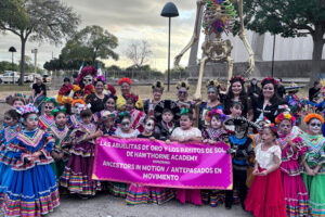 Children and adults dressed in colorful ballet folklorico outfits and dia de los muertos face paint, gather outdoors holding a banner in front of a tall skeleton decoration.