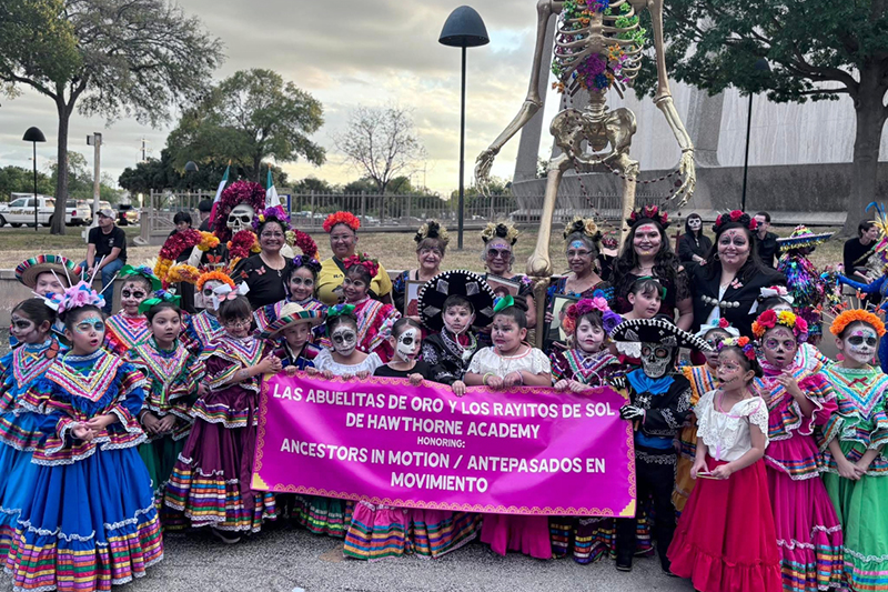 Children and adults dressed in colorful ballet folklorico outfits and dia de los muertos face paint, gather outdoors holding a banner in front of a tall skeleton decoration.