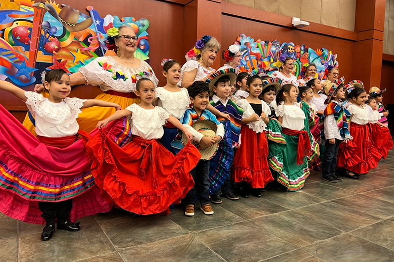 A line of children and older adults dressed in colorful ballet folklorico outfits pose indoors, holding out vibrant skirts in front of a bright mural backdrop.