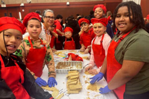 A group of children and an adult wearing matching red chef hats and aprons gather around a table indoors while preparing tamales together.