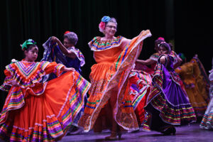 A group of performers on a stage wear bright, ballet folklorico dresses and hold out the flowing skirts while dancing.