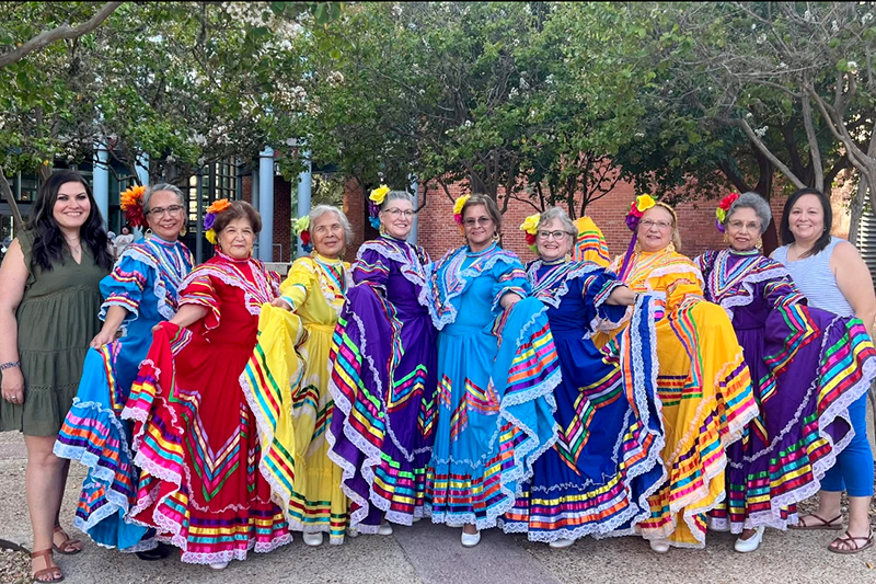 Las Abuelitas do Oro and their sponsors stand outdoors in a line, dressed in brightly colored ballet folklorico dresses with patterned trim, holding out their skirts in a pose beneath leafy trees near a building.