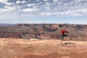 Ruth balances on one leg near the edge of a vast red-rock canyon landscape under a partly cloudy sky.