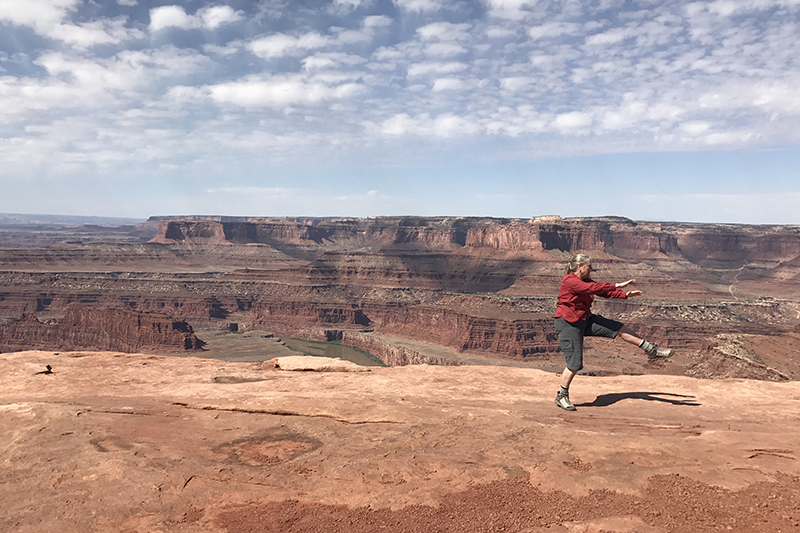 Ruth balances on one leg near the edge of a vast red-rock canyon landscape under a partly cloudy sky.