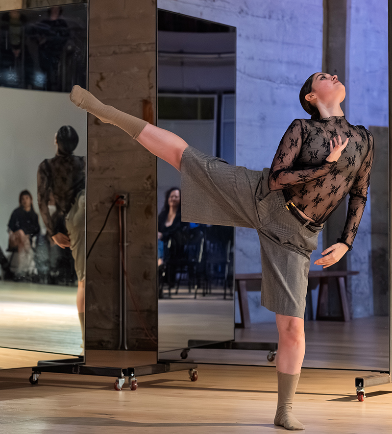 A dancer balances on one leg while extending the other horizontally and arching the torso beside mirrored panels in a studio space.