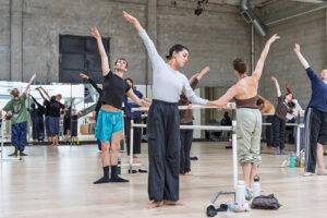 Dancers in a ballet class stand in first position with raised arms around barres in a spacious studio with mirrored walls and industrial-style architecture.