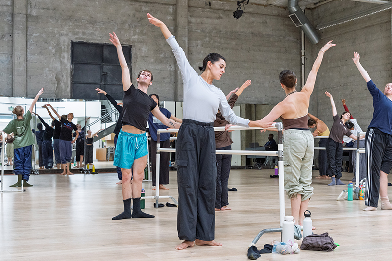 Dancers in a ballet class stand in first position with raised arms around barres in a spacious studio with mirrored walls and industrial-style architecture.