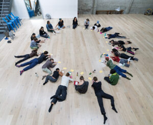 A large group of people lie and sit in a wide circle on a wooden studio floor, writing on colorful papers during a collaborative activity.