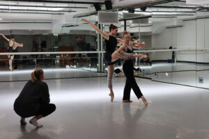 Two dancers en pointe practice a partnered movement in a mirrored studio while a third person crouches nearby observing, with reflections and ballet barres visible along the walls.