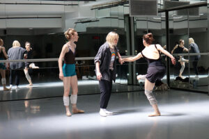 In a mirrored dance studio, three individuals stand together as one performs a lifted, turning movement while another holds the performer’s hand and a third observes nearby.