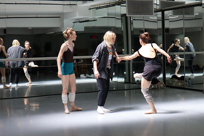In a mirrored dance studio, three individuals stand together as one performs a lifted, turning movement while another holds the performer’s hand and a third observes nearby.