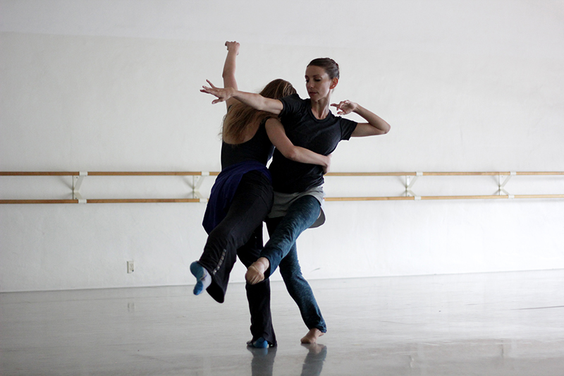 Two people perform a contemporary dance duet in a bright studio, intertwining their bodies in a dynamic pose near a wall-length ballet barre.