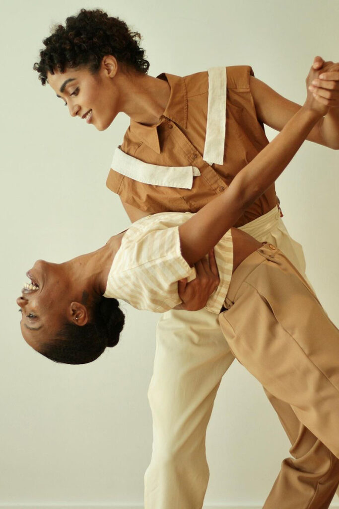 Two people pose in a studio against a plain background, with one participant supporting and dipping the other in a graceful dance-like movement while both wear coordinated neutral-toned outfits.