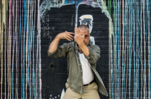 Joe stands in front of a black brick wall covered in streaked graffiti, holding arms in a dynamic, blurred motion.