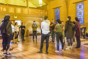 A group of people stand in a circle inside a warmly lit room with wooden floors and stained glass windows, participating in a group activity or workshop.