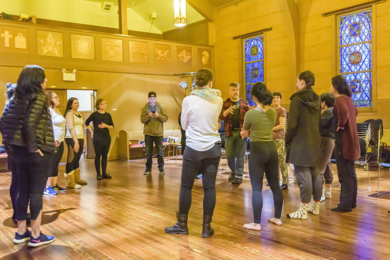 A group of people stand in a circle inside a warmly lit room with wooden floors and stained glass windows, participating in a group activity or workshop.