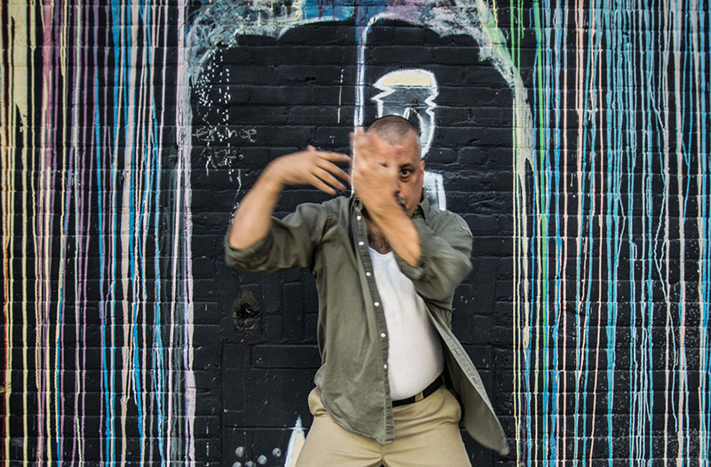 Joe stands in front of a black brick wall covered in streaked graffiti, holding arms in a dynamic, blurred motion.
