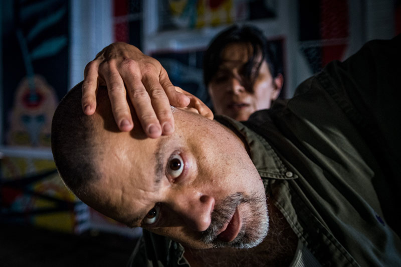 Joe's face is in the foreground with someone's hand reaching over his forehead. Another person is in the background, darkly lit.